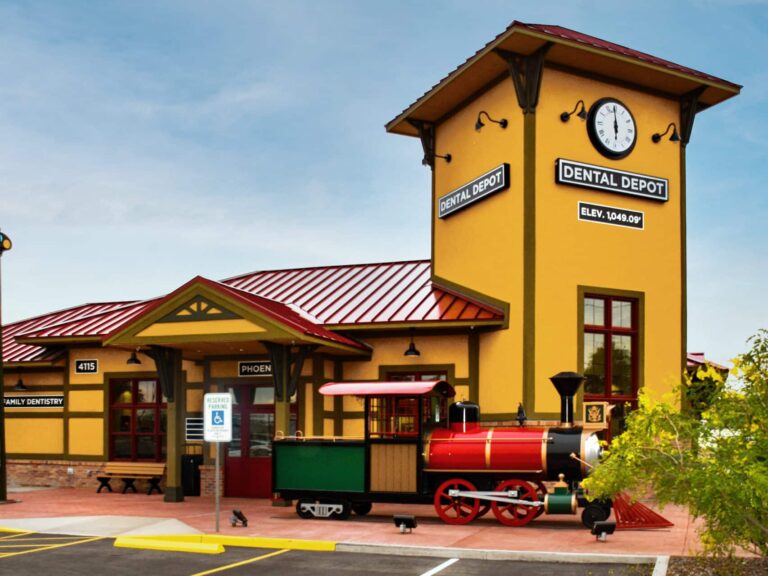 Exterior view of Dental Depot's  West Phoenix dentist office with a clock tower, model train, yellow walls, and a red roof.