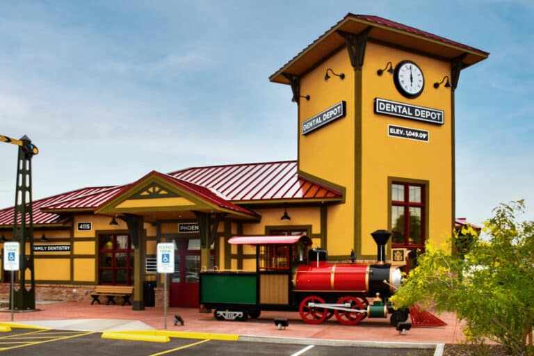 Exterior view of the Dental Depot dentist office in West Phoenix with a clock tower, model train, red roof, and yellow exterior.