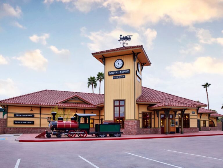 Exterior view of Dental Depot's dentist office in Mesa, AZ with a clock tower, yellow exterior, red roof, and model train in front