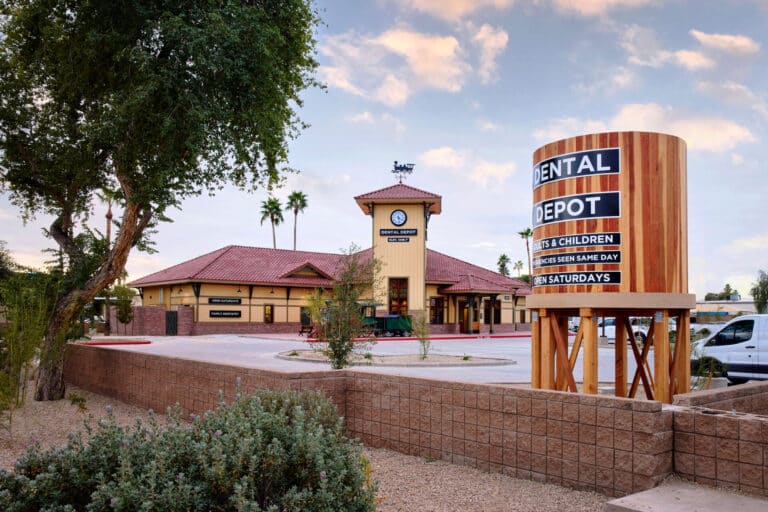 Exterior view of Dental Depot's dentist office in Mesa, AZ with a clock tower, yellow exterior, red roof, water tower, and model train in front