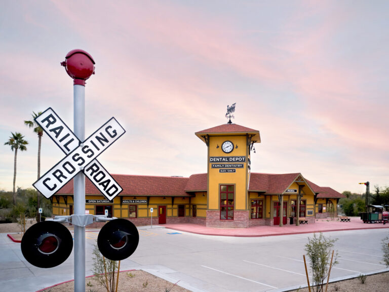 Exterior view of Dental Depot's dentist office in Peoria, AZ with a model train, yellow exterior, railroad crossing sign, and clock tower