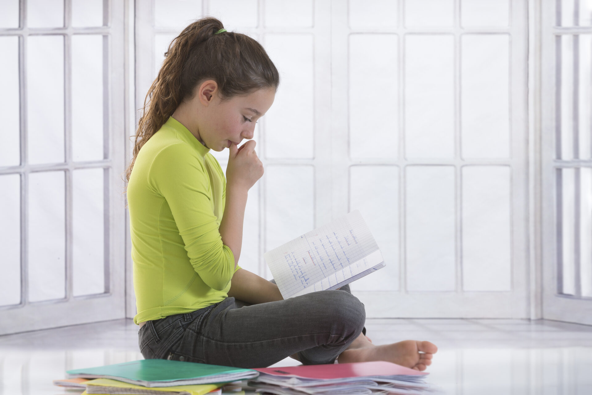 Girl in a lime green shirt and grey sweats sits while reading a book and sucking her thumb.