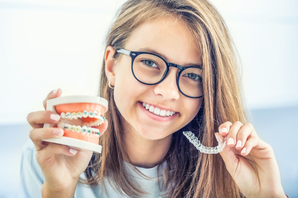 Young lady in glasses holds up models of Invisalign and braces to compare them.