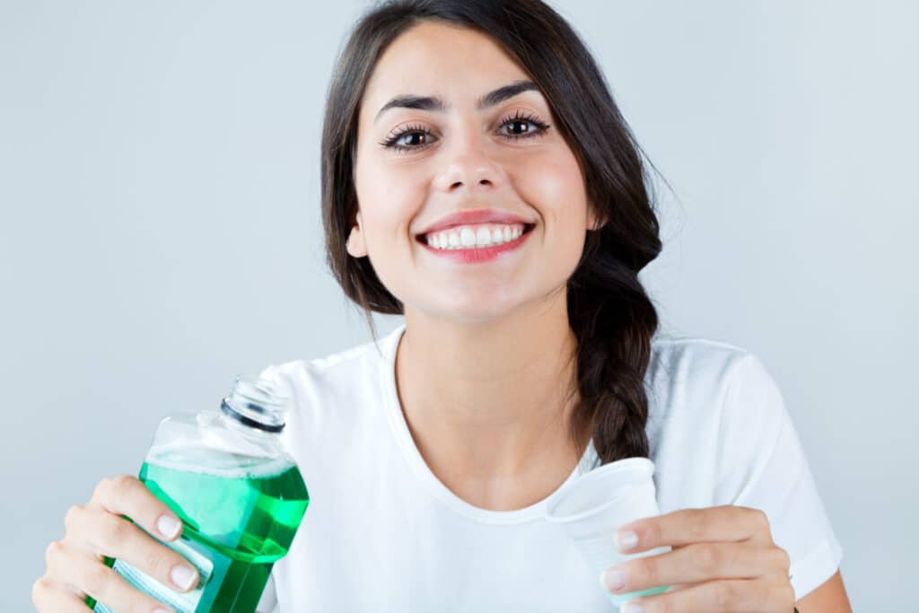 Portrait of pretty brunette girl using green mouthwash and smiling for the camera, isolated on white background.