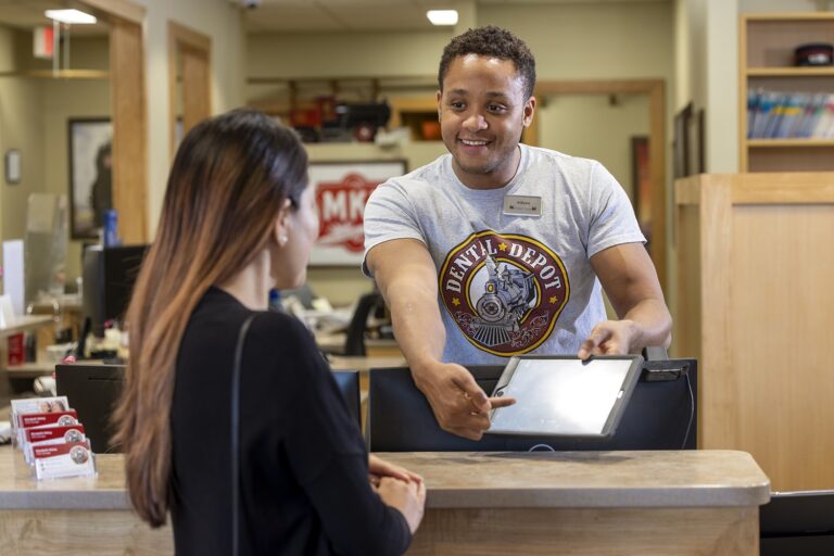 An attendant at Dental Depot's front desk in a dental office holds out a clipboard while smiling and helping a patient.