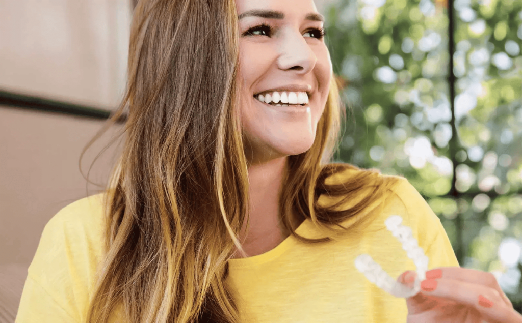 A woman in a yellow shirt smiles while holding Invisalign aligners.