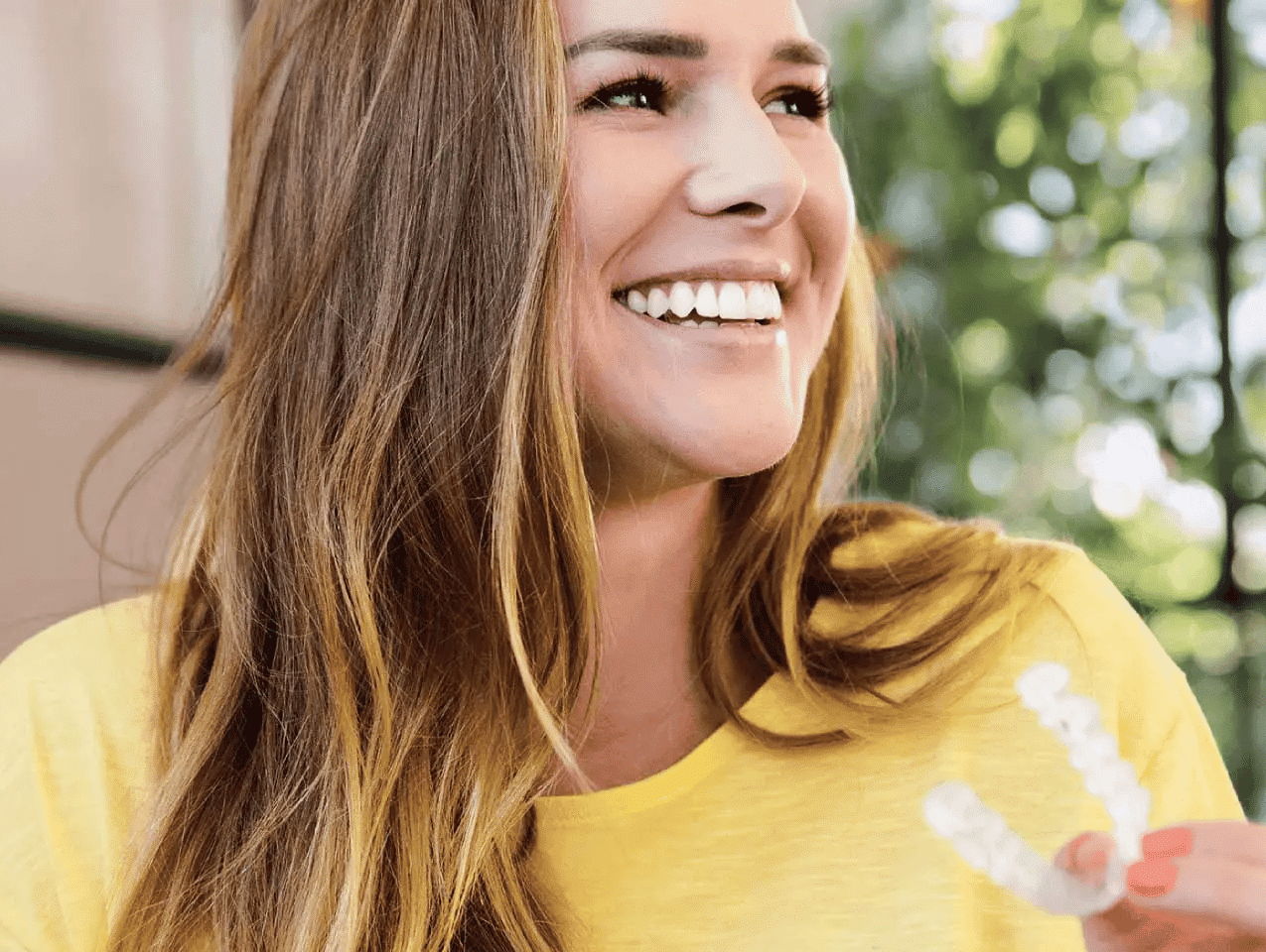 A woman in a yellow shirt smiles while holding Invisalign aligners.